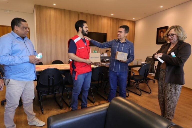 People in an office exchanging small boxes; one man in a red vest hands a box to another man while a woman holds a bottle and a tube nearby.