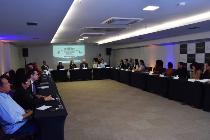 Group of professionals seated around a U-shaped conference table in a meeting room, with a projector screen at the front displaying a presentation.