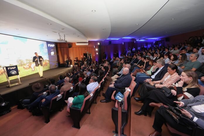 Speaker on stage delivering a talk to a large audience in a tiered auditorium, with a bright screen and FIAP branding on the podium.