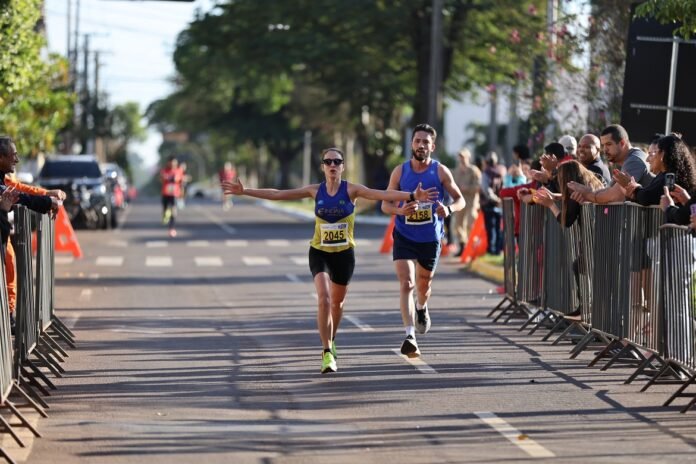 Two runners finish a street race with arms outstretched, crowds cheering behind metal barriers on a tree-lined road.