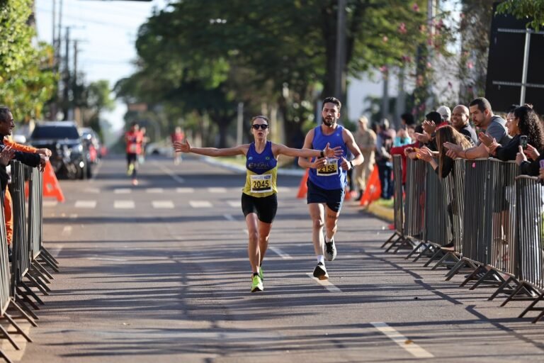 Two runners finish a street race with arms outstretched, crowds cheering behind metal barriers on a tree-lined road.