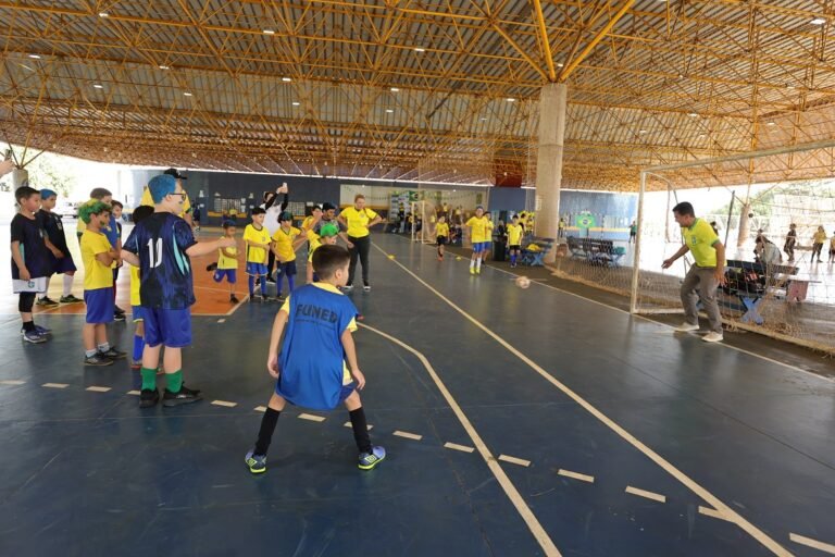Youth soccer practice inside a covered gym: kids in blue and yellow uniforms watch as a coach kicks toward the goal.
