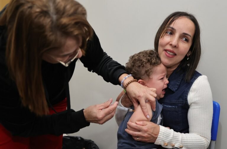 Toddler crying during vaccination as a caregiver administers a shot while a mother holds him for support.