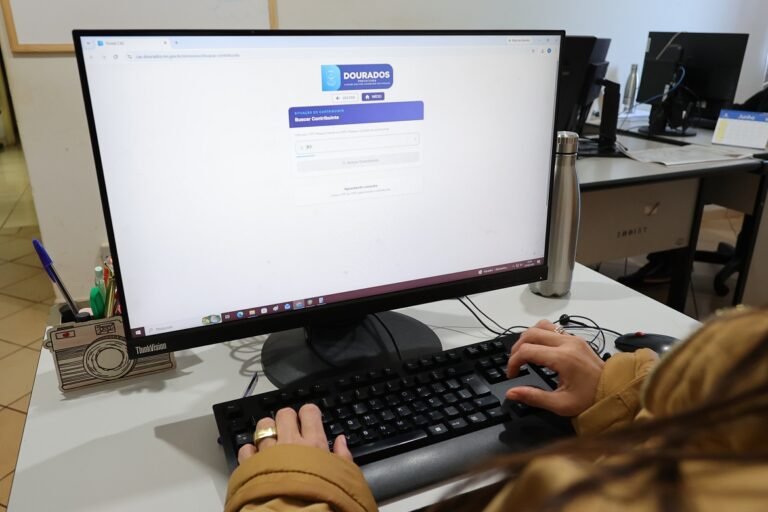 Person typing on a keyboard at a desk with a large monitor showing a government site form labeled 'Buscar Contribuinte' (Search Contributor).