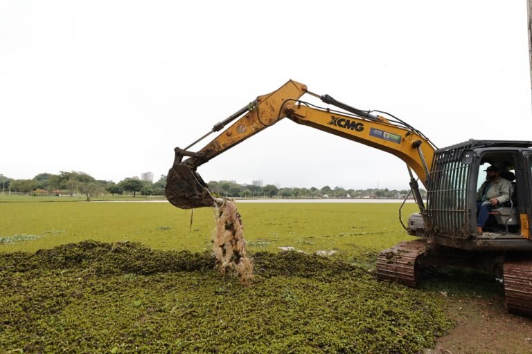 Yellow excavator (XCMG) on a muddy field pitches a bucket of mud and water, with the operator visible in the cab at right edge of the frame.