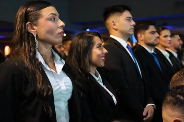 Group of young professionals in business attire standing in a dim conference hall, facing left and listening intently to a speaker.