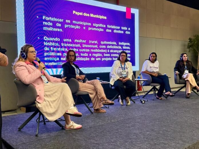 Five women panelists seated on a stage; one speaks into a microphone, with a purple presentation slide behind them reading 'Papel dos Municípios'.