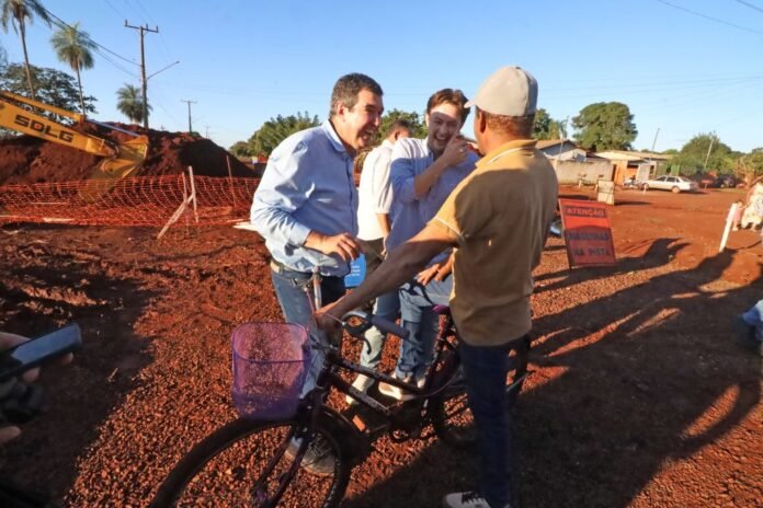 Three men chat and smile beside a bicycle with a purple basket on a dusty construction site, with orange safety fencing and a backhoe in the background.