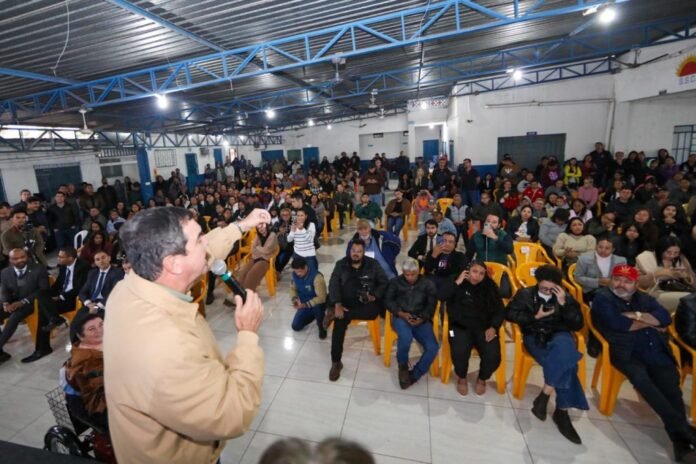 Speaker in beige jacket addresses a large audience seated on yellow chairs in a gym-like hall with blue beams and bright lighting.