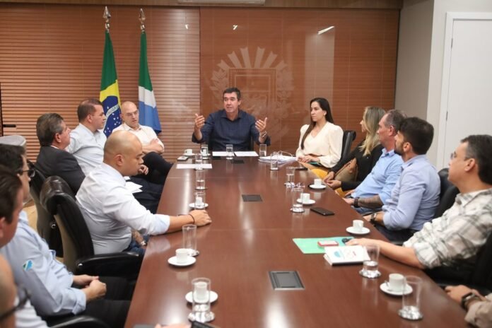 Group of professionals seated around a long conference table in a meeting room, with two flags in the background (Brazil and another) and beverages on the table.