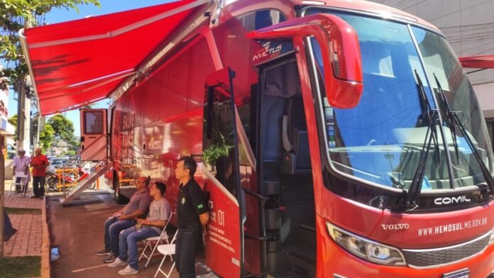 Red mobile service bus with its door open and stairs down, awning extended, as people sit on folding chairs beside it on a sunny street.