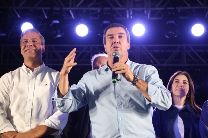 Center man in a light blue shirt speaks into a microphone on a stage, flanked by two supporters under bright stage lights.