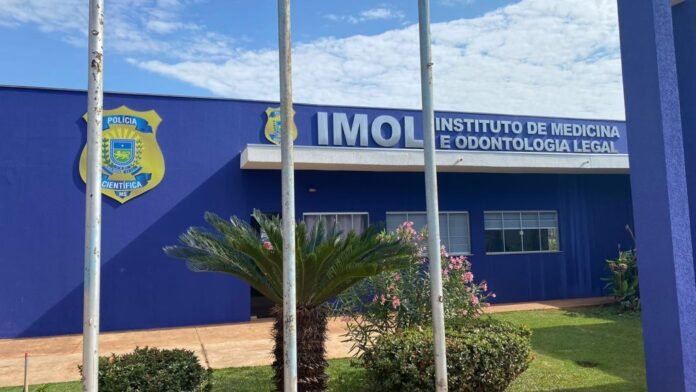 Blue building with a sign reading 'INSTITUTO DE MEDICINA E ODONTOLOGIA LEGAL' and a police shield on the left, with palm plants and flowering shrubs in front.