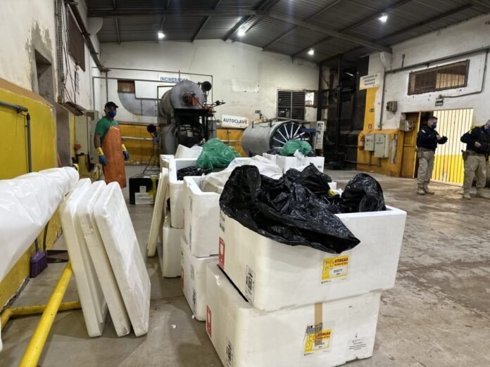 Industrial waste processing area with large white foam blocks, green and black bags, and PPE-clad workers in a high-ceiling facility.