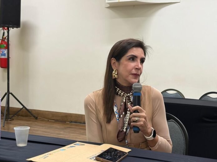 Woman seated at a table speaking into a handheld microphone during a panel or interview, with a beige blouse and polka-dotted scarf; a tablet and a paper folder are on the table beside a plastic cup.