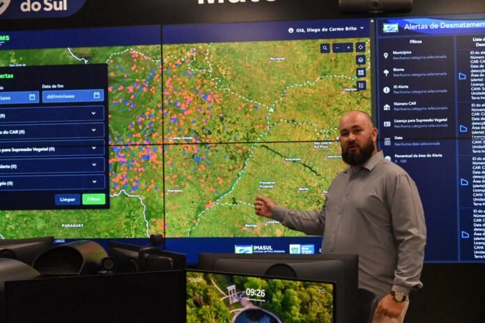 Man with a beard in a gray shirt stands beside a large wall of screens showing a deforestation alert map and controls in Portuguese, gesturing at the map.