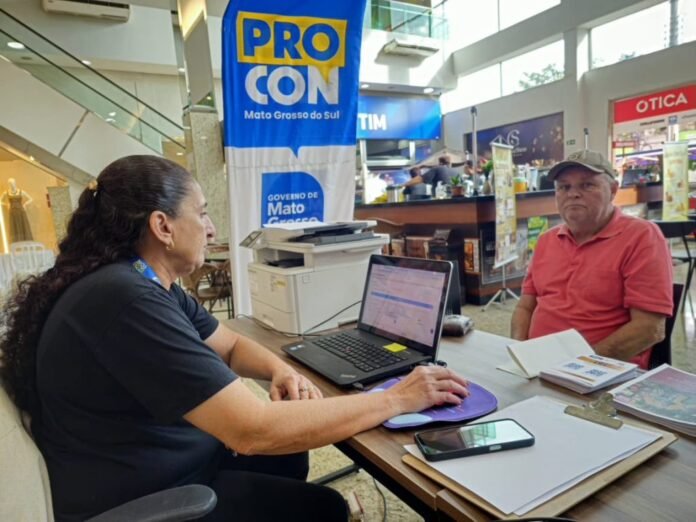 A woman speaks with a man across a desk at a public event stall with a PRO CON banner in the background and a laptop on the table.