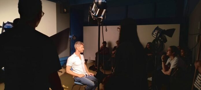 Man in a white shirt sits on a chair in a professional studio as crew film an interview under bright studio lights.