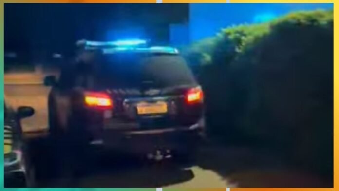 Rear view of a black SUV with blue flashing police lights on top, driving away at night along a hedge-lined street.
