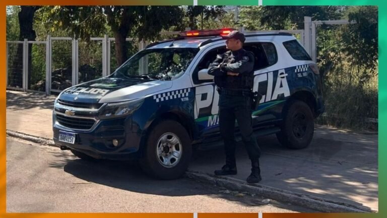 Policial em uniforme com braços cruzados ao lado de uma viatura da polícia azul e branca, estacionada na calçada.