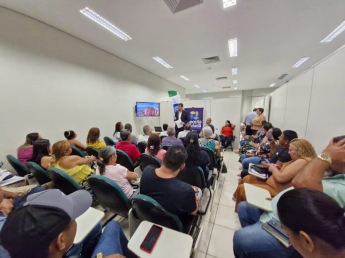A man gives a talk to a large audience in a bright conference room; attendees sit in rows, with a screen and banners behind the speaker.