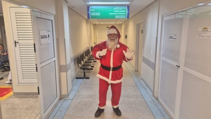 Man in a Santa Claus costume standing in a hospital corridor, waving with both hands, with chairs along the wall and closed doors nearby.