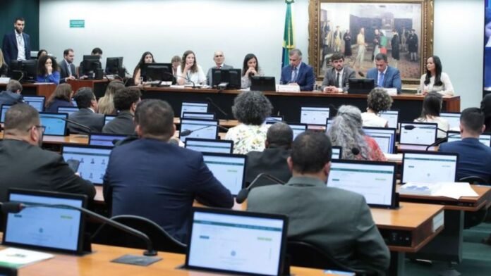 Panel of speakers at a formal meeting in a conference room, attendees in the audience using laptops beforehand forming rows of screens at desks.