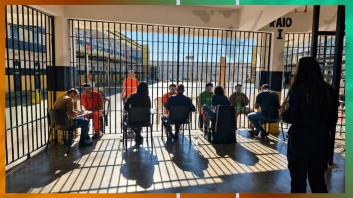 Inmates seated in chairs behind tall bars inside a prison yard, with a guard in orange standing nearby and shadows crossing the floor.