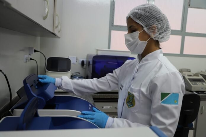 Lab technician in PPE (mask, hairnet, gloves) uses a blue centrifuge on a lab bench.