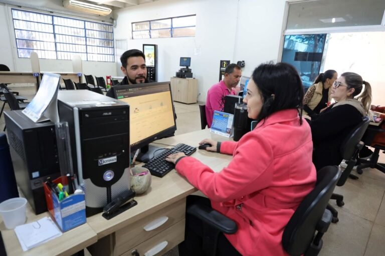 Woman in a pink jacket typing at a computer at a busy service desk in an open office setting.