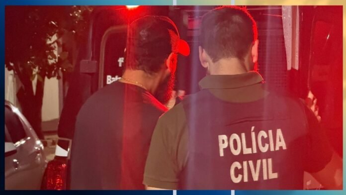 Two men are seen from behind at night, one wearing a Polícia Civil vest, near a vehicle with red lighting in the background.