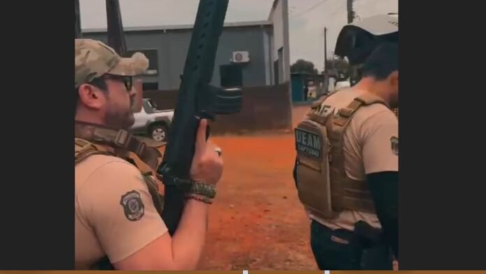 Two law-enforcement officers in tan uniforms and tactical vests, one holding a rifle outdoors on an orange dirt area coolly readying his weapon.