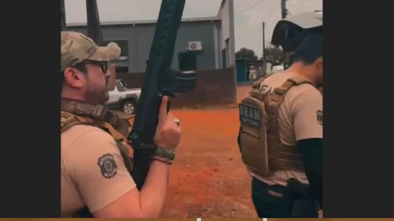 Two law-enforcement officers in tan uniforms and tactical vests, one holding a rifle outdoors on an orange dirt area coolly readying his weapon.