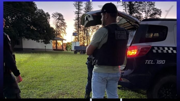 Police officers near an open car trunk at dusk, one wearing a tactical vest and jeans, in a grassy area.