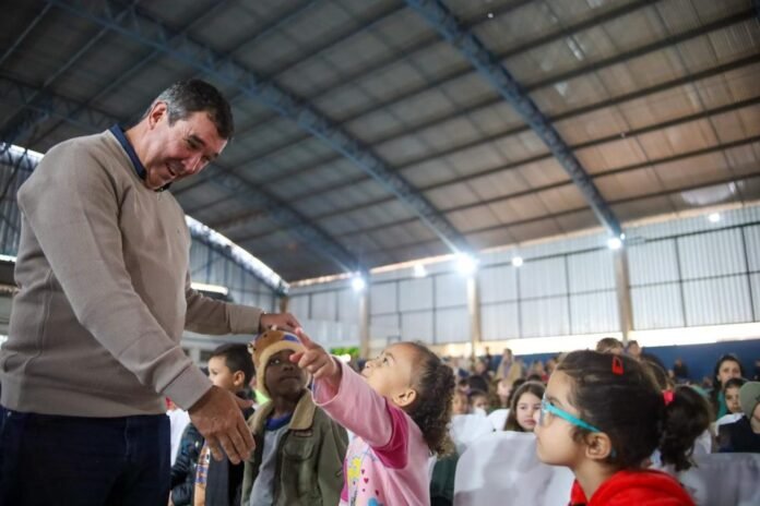 A man in a beige sweater greets a young girl reaching out to shake his hand at an indoor community event with many children seated in rows in the background.