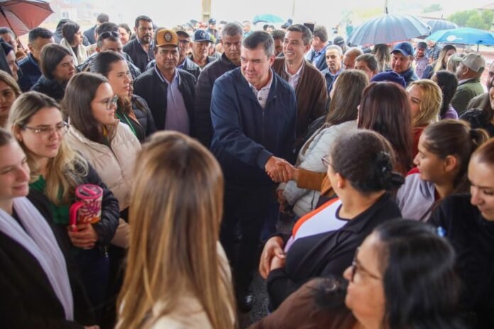 Man in a dark jacket shakes hands with a woman in a crowd at an outdoor event in the rain; numerous onlookers surround them under umbrellas.