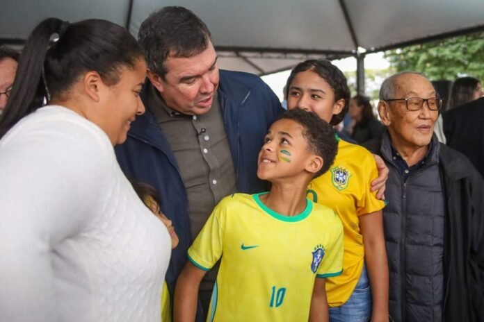 Boy in a yellow Brazil jersey with green face paint looks up at a man in a navy jacket, while others smile nearby under a canopy.