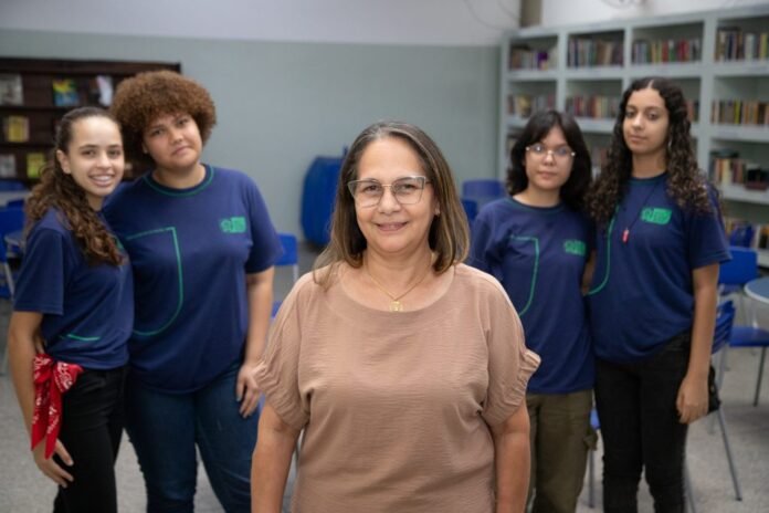 Smiling middle-aged woman in a beige top standing in front of four students in blue shirts in a library setting.