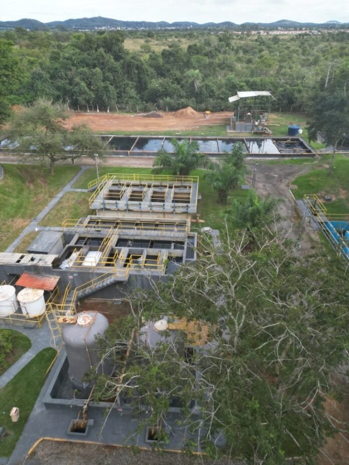 Aerial view of a wastewater treatment plant with concrete basins, yellow safety railings, and surrounding trees and greenery.