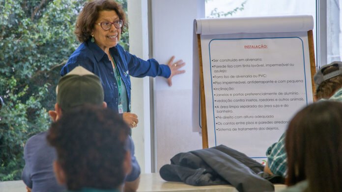Woman presenter in blue shirt standing beside a flip chart, speaking to a small audience in a bright room.