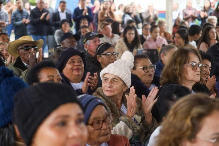 Diverse crowd at an outdoor event, people clapping, with a woman in a white knit hat at center of the frame.