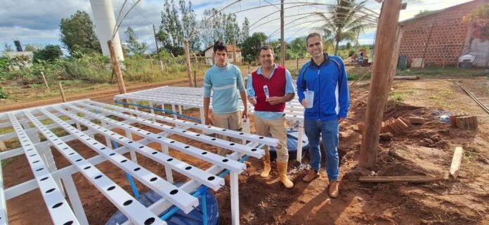 Three workers stand beside a white perforated metal frame on a dirt construction site, with a blue water tank nearby and a brick building in the background.