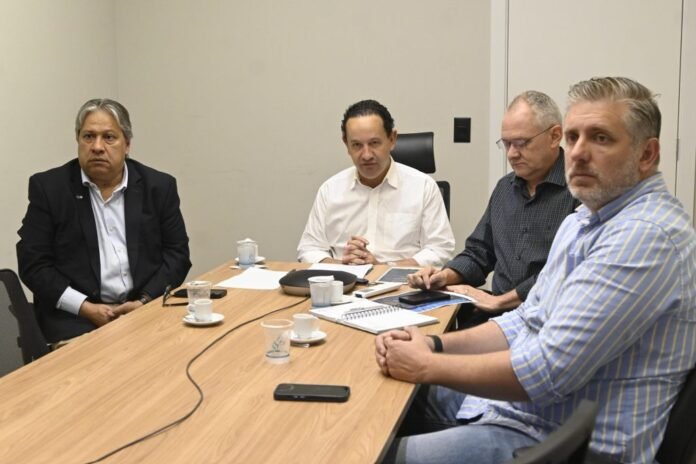 Four men sit around a wooden conference table in a meeting room, looking attentive as they discuss documents and devices on the table.