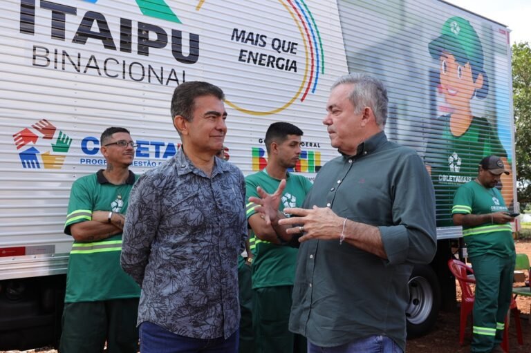 Two men in casual shirts chatting in front of a large Itaipu Binacional truck, with workers in green uniforms behind them.
