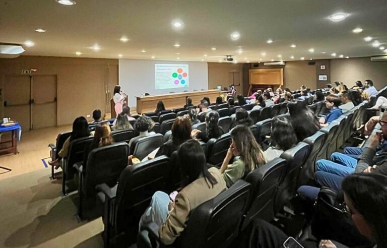 Speaker at a podium presenting to a large audience in an auditorium with a projected slide on the screen above the desk area.
