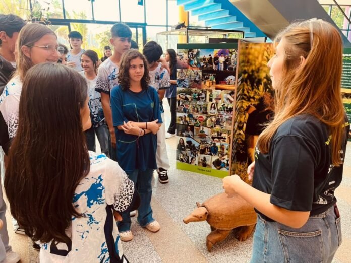 Group of students listening to a presenter beside a collage poster at an indoor exhibition, with a carved wooden boar on the floor nearby.