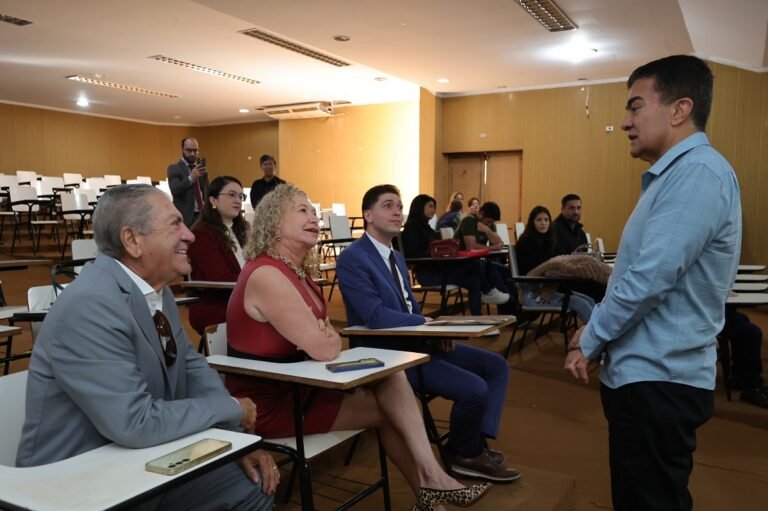 Man stands at the front speaking to a seated audience in a classroom lecture hall.