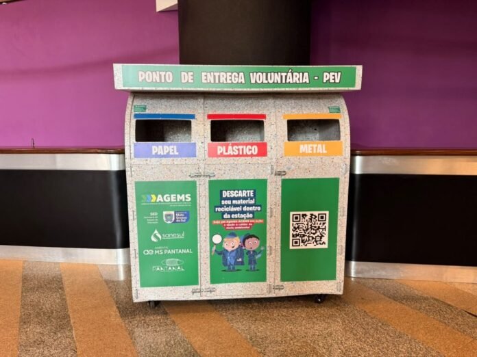 Recycling station with three labeled bins for Paper, Plastic, and Metal under a sign reading 'Ponto de entrega voluntária - PEV', against a purple wall.