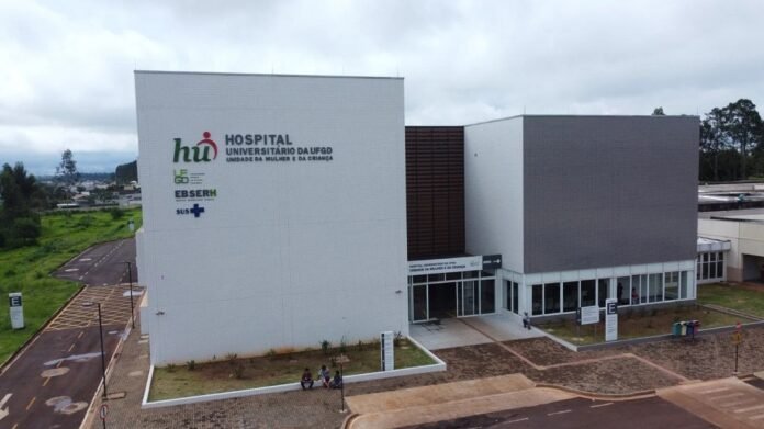 Exterior view of a modern hospital building with a large white facade and signage reading 'Hospital Universitário da UF..., Unidade da Mulher e da Criança' in Portuguese, plus logos on the left.