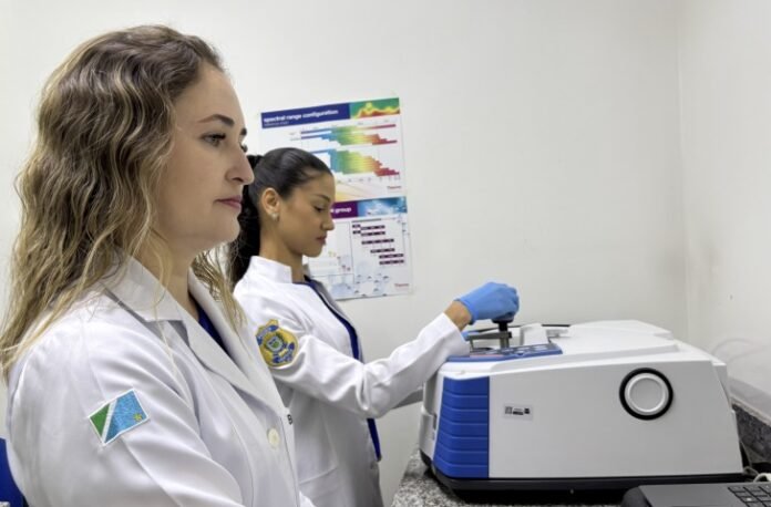 Two female scientists in white coats operate a laboratory instrument in a clean lab setting.